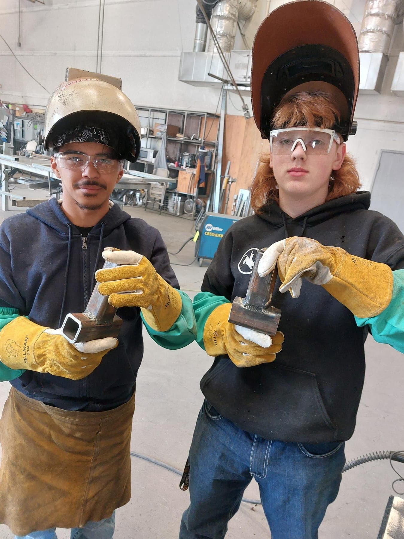 Two students in welding helmets holding their metalwork